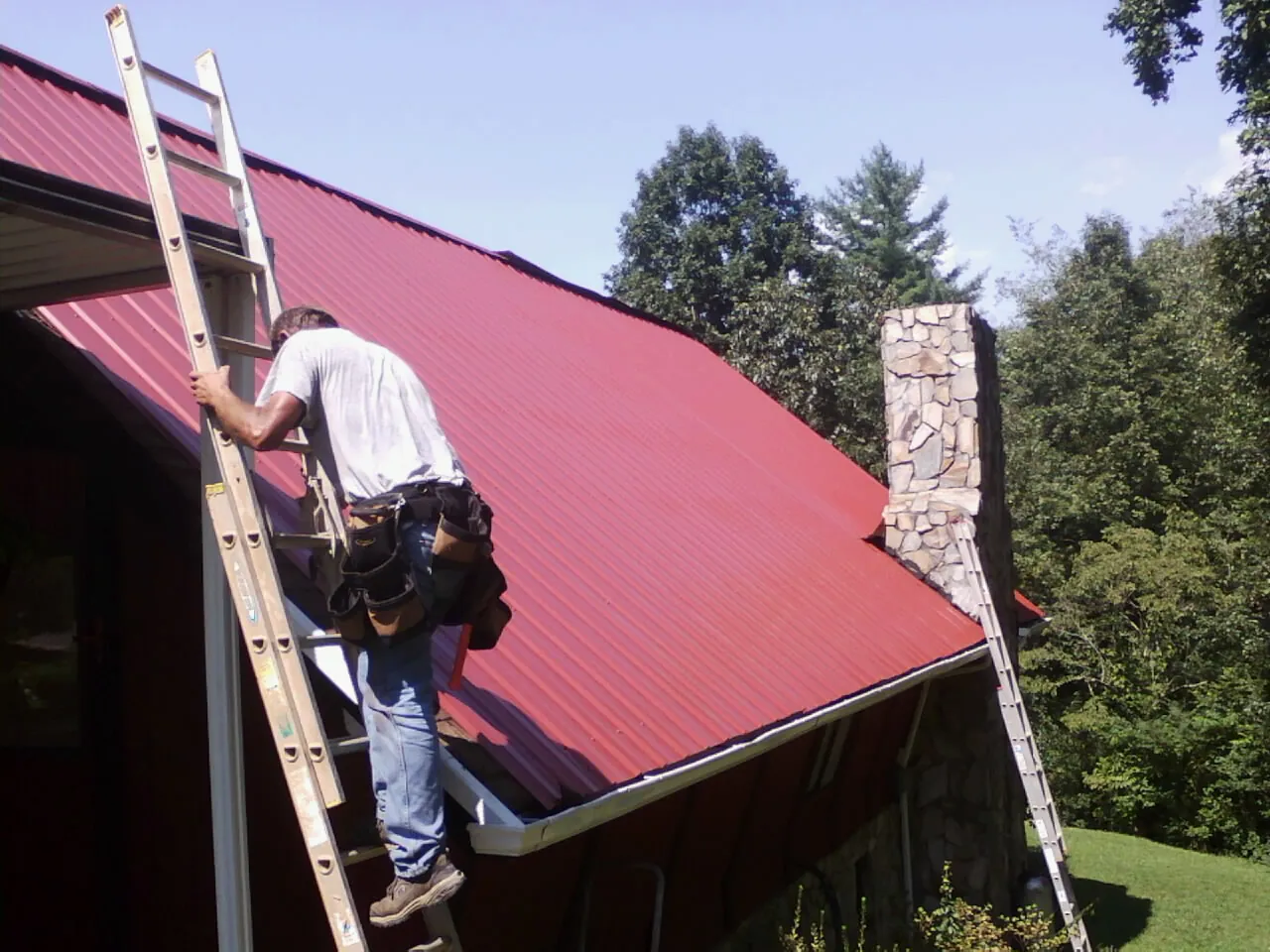 Roofer on a ladder beside a red metal roof and stone chimney — Soffit & Fascia Repair by Roofing Brothers in Spraggs